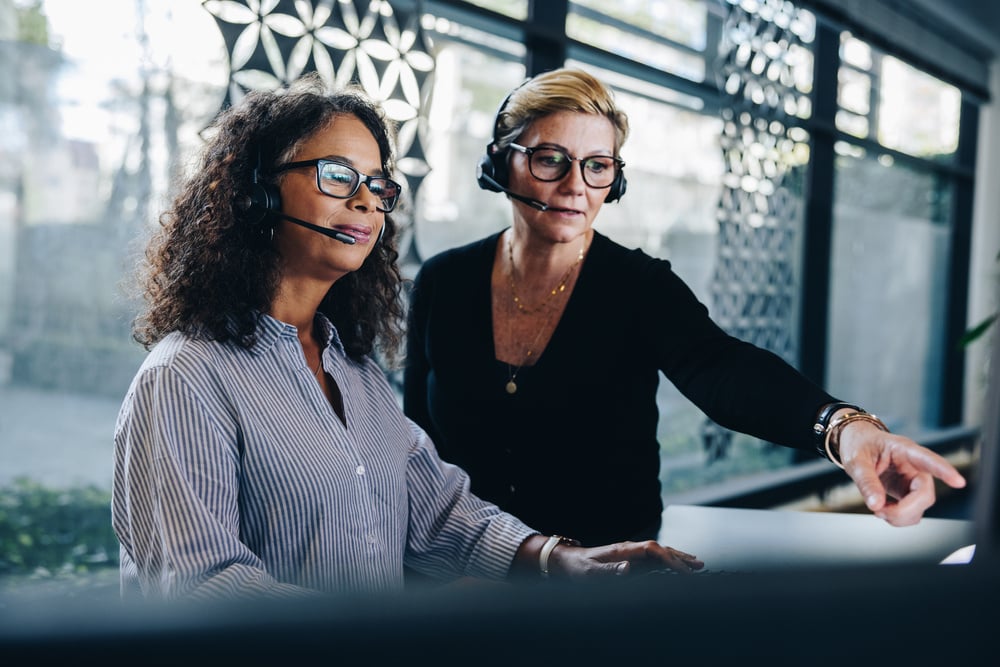 Two women with headsets on, focused on a computer screen together